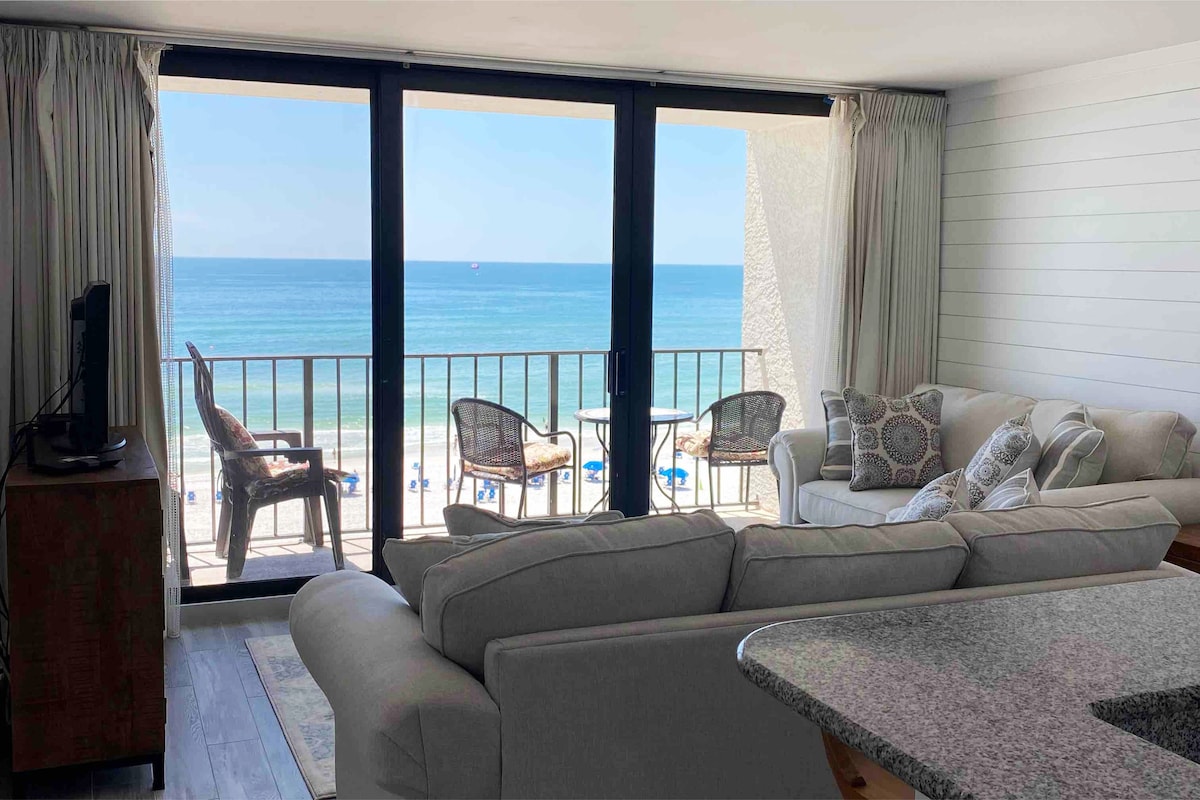 A modern living area is highlighted by large glass doors that offer a view of the ocean. A light-colored sofa with decorative pillows is positioned beside a wooden TV stand. Natural light fills the space, creating a bright and airy atmosphere.