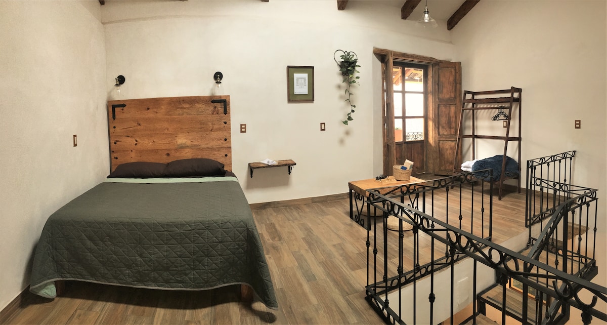 A spacious bedroom features a wooden bed frame with a textured blanket, complemented by a side table and wall-mounted lamps. A decorative plant hangs near a window that opens to a balcony, while a staircase leads to the lower level. A simple seating area is present.