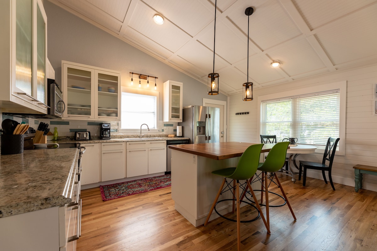 An open-concept kitchen is depicted, featuring a central island with bar seating and sleek green stools. Light wood flooring complements the granite countertops and white cabinetry. Large windows provide natural light, while various kitchen appliances are neatly arranged along the countertops.