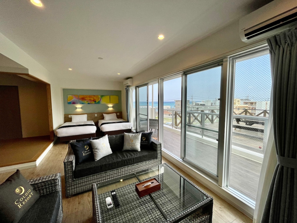 A spacious room features two beds, large windows providing natural light, and a view of the balcony. Modern furnishings include a comfortable seating area with a glass table and wicker chairs, creating an inviting space for relaxation and enjoyment.