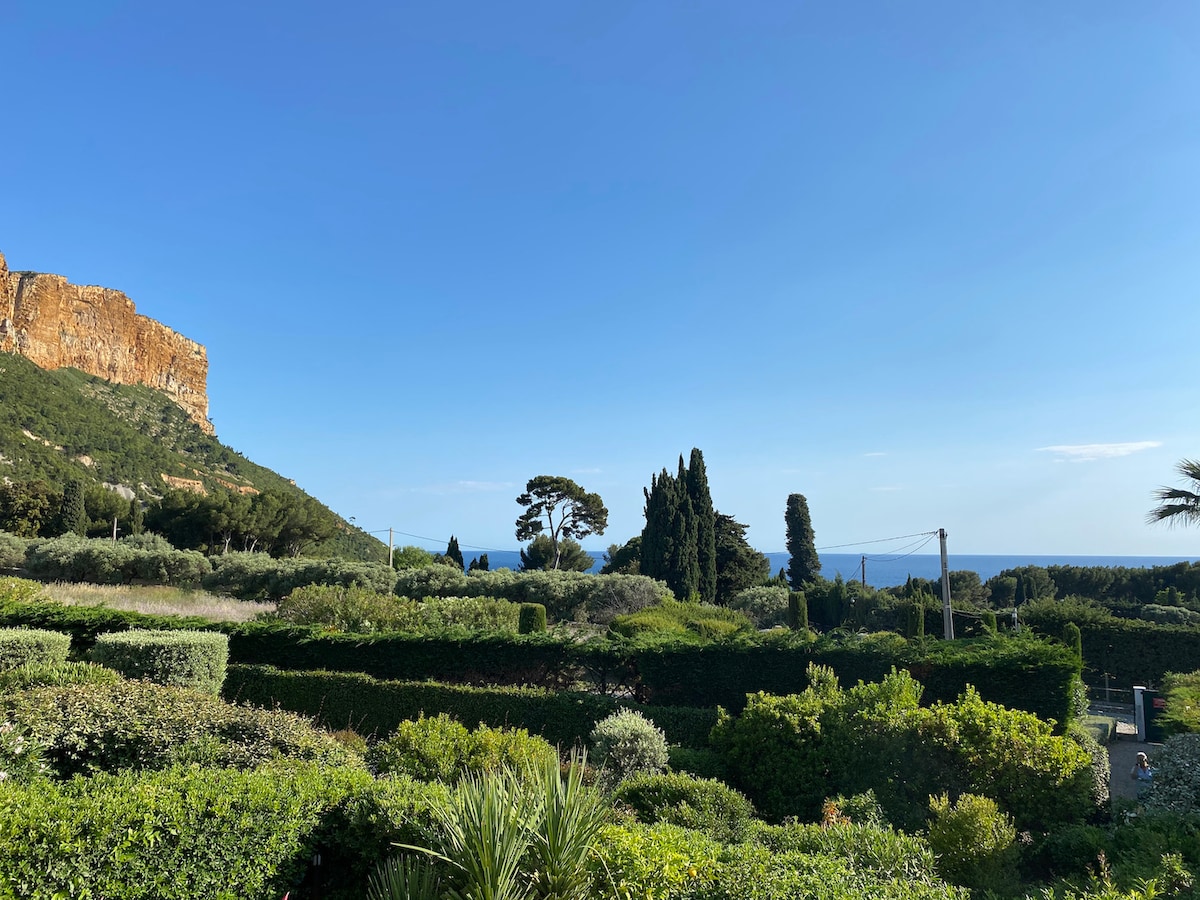 A scenic view features lush greenery and hedges, contrasting with the blue sky and distant sea. The rocky cliff is visible in the background, with tall trees adding depth. The serene landscape showcases the natural beauty surrounding the residence.