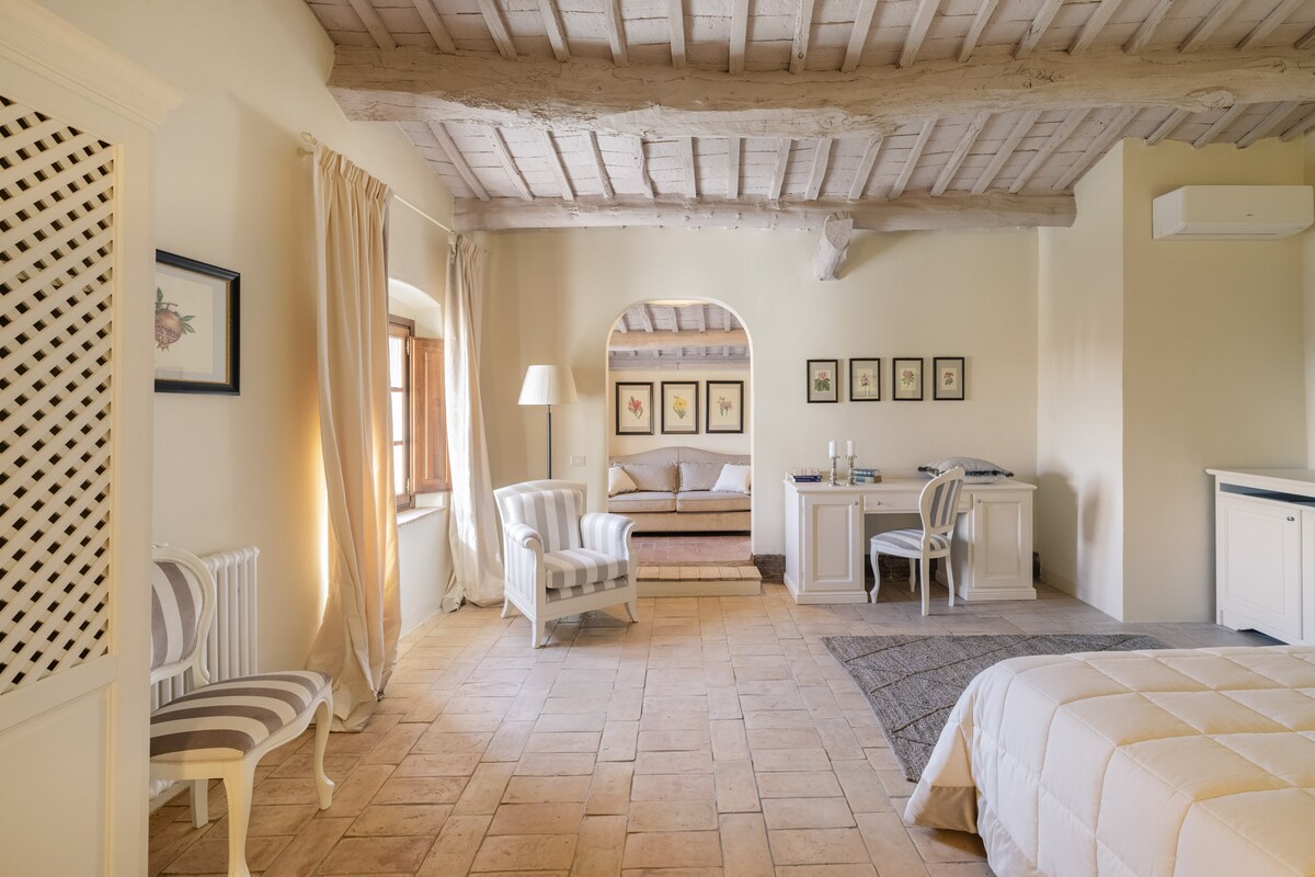 A spacious bedroom features a combination of natural light and soft neutral tones. Elegant wooden beams line the ceiling above. Two striped armchairs are positioned beside a lamp, while a cozy bedding area is situated toward the back, complemented by a desk and a small seating area.