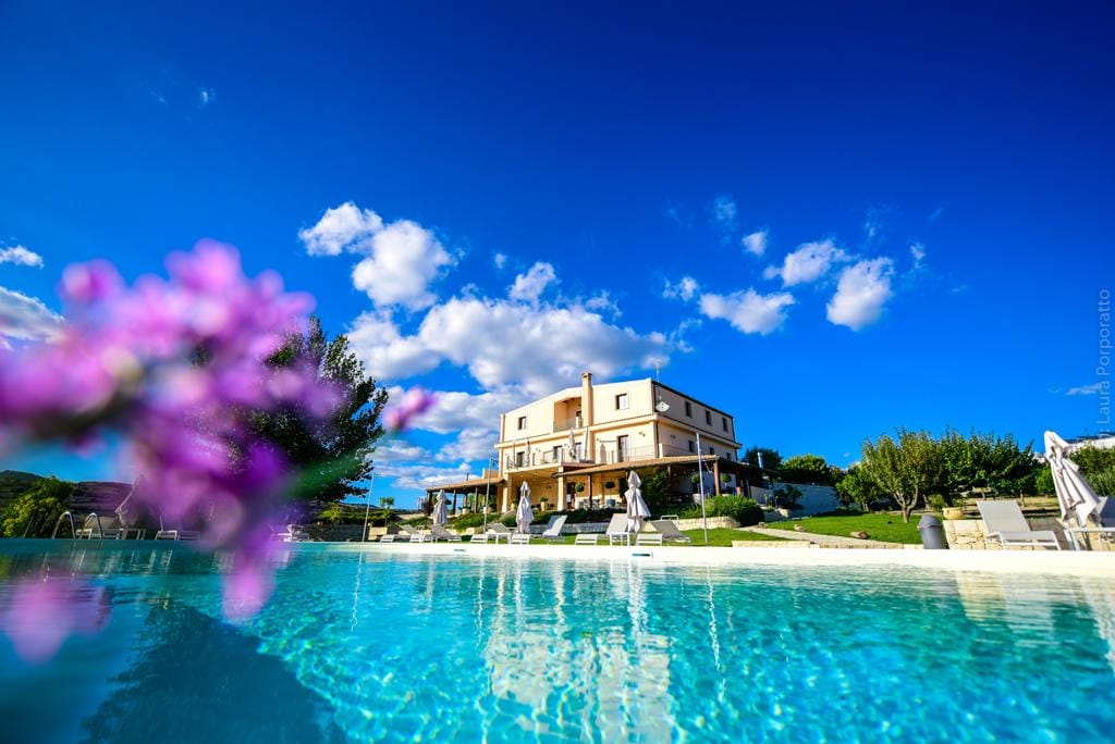 The inviting swimming pool reflects a clear blue sky flecked with white clouds. In the background, a spacious villa with multiple outdoor seating areas is framed by lush greenery. A portion of vibrant flowers adds a touch of color to the forefront.