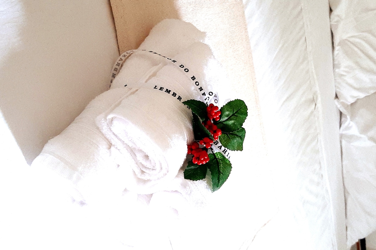 Neatly arranged white towels are presented with an artificial floral decoration featuring small red berries. A clean and neutral bed linen is visible in the background, contributing to a fresh look.