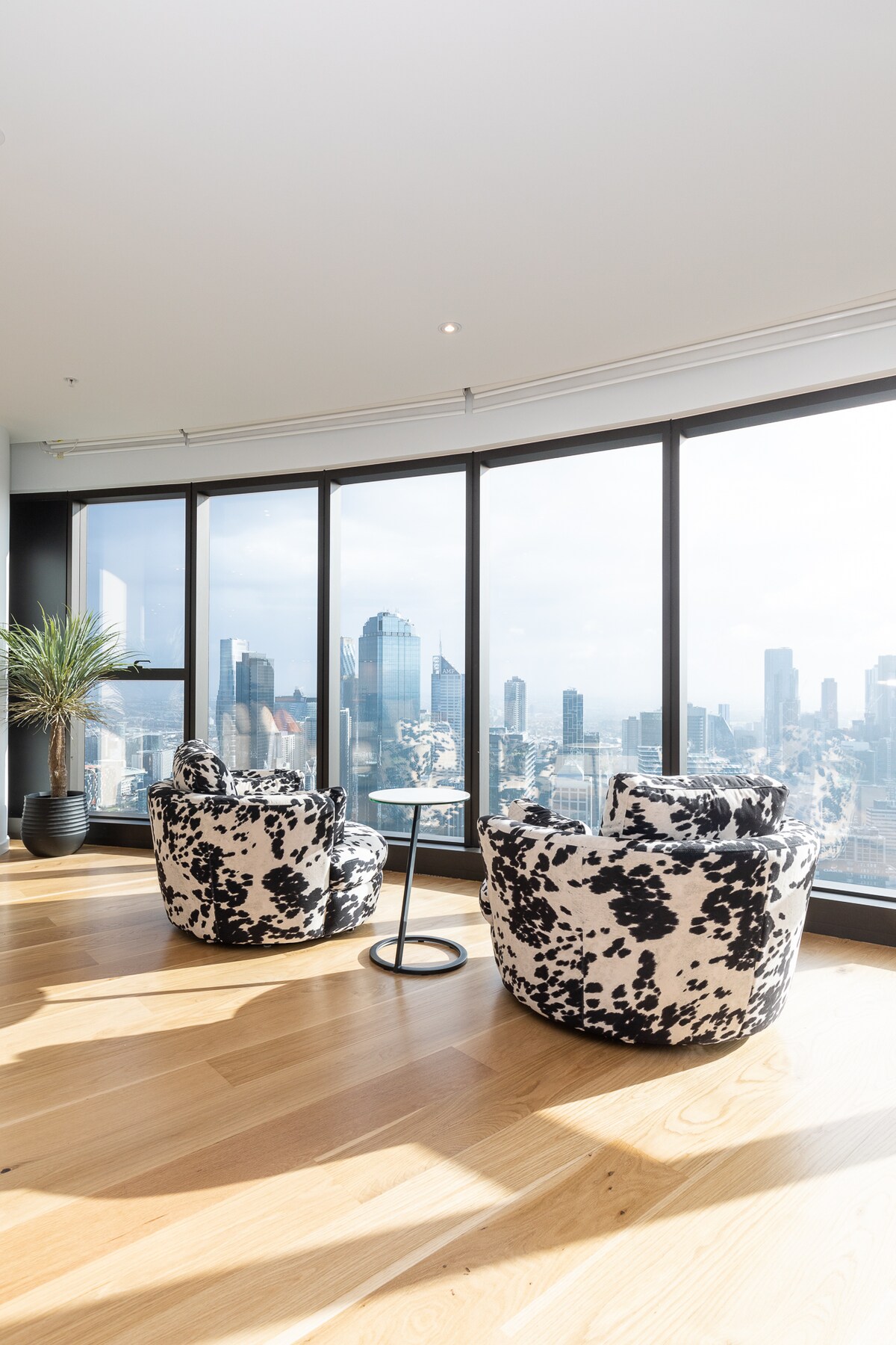 Two armchairs with a monochrome pattern are positioned next to a small, round table. Large floor-to-ceiling windows offer expansive views of the Melbourne skyline, allowing natural light to fill the space, while a potted plant adds a touch of greenery.