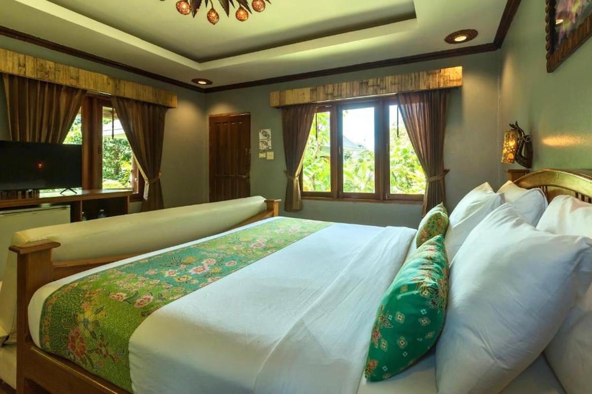 A spacious bedroom features wooden floors and Thai-style decorations. Two large beds are adorned with colorful pillows. Natural light filters through the curtains, illuminating the cozy seating area and a TV unit. Lush greenery is visible through the windows, enhancing the room's serene ambiance.
