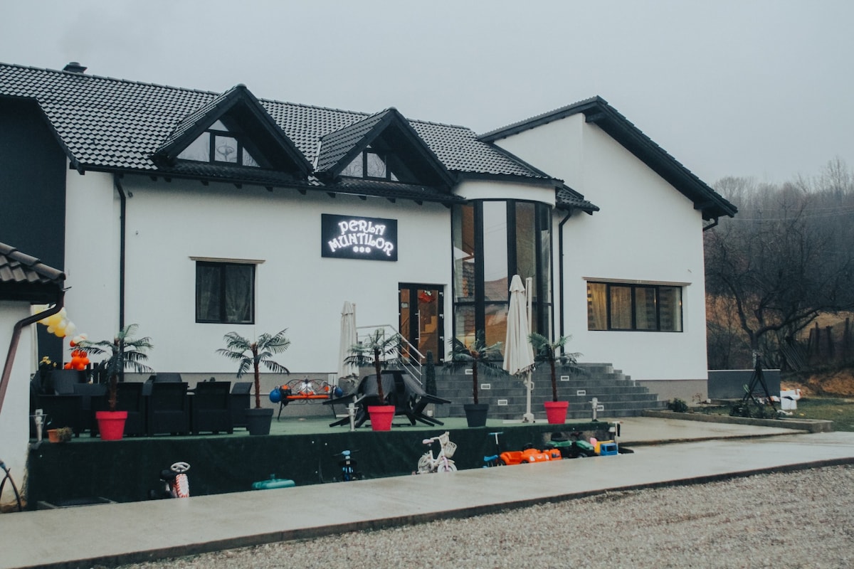 The exterior of a spacious building is shown, featuring a mix of modern and traditional architectural elements. The entrance includes steps leading to a welcoming porch, surrounded by potted plants. Outdoor seating areas are visible, offering inviting spaces for relaxation.