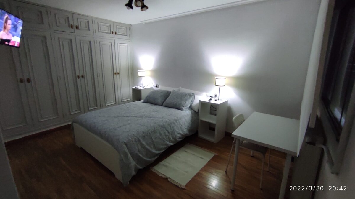 A private bedroom features a spacious bed with soft linens, complemented by two bedside lamps. A television is mounted on the wall opposite the bed. A light-colored desk and a small chair are positioned beside a large wardrobe, creating an organized living space.