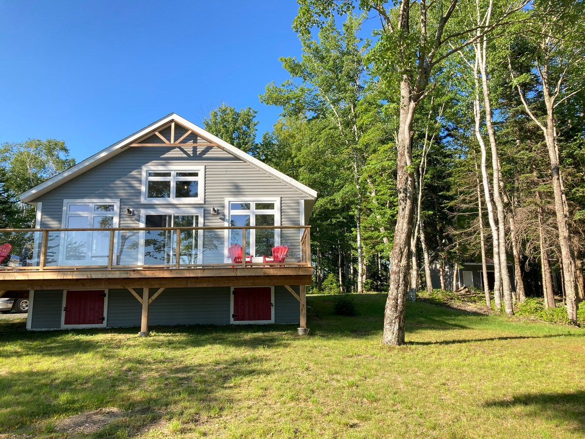 The exterior of a modern gray cottage is highlighted, featuring a spacious deck with red patio chairs. Large windows enhance natural light, and surrounding trees provide a serene setting. A well-maintained lawn leads to a wooded area in the background.
