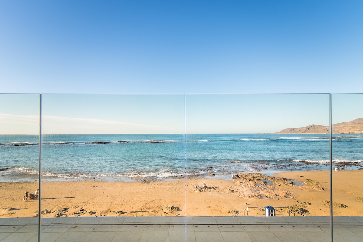 A clear view of the beach and ocean is presented, with a sandy shore extending along the coastline. The calm water reflects the blue sky, while gentle waves break on the rocky areas. The scene is framed by a transparent glass barrier, enhancing the coastal perspective.