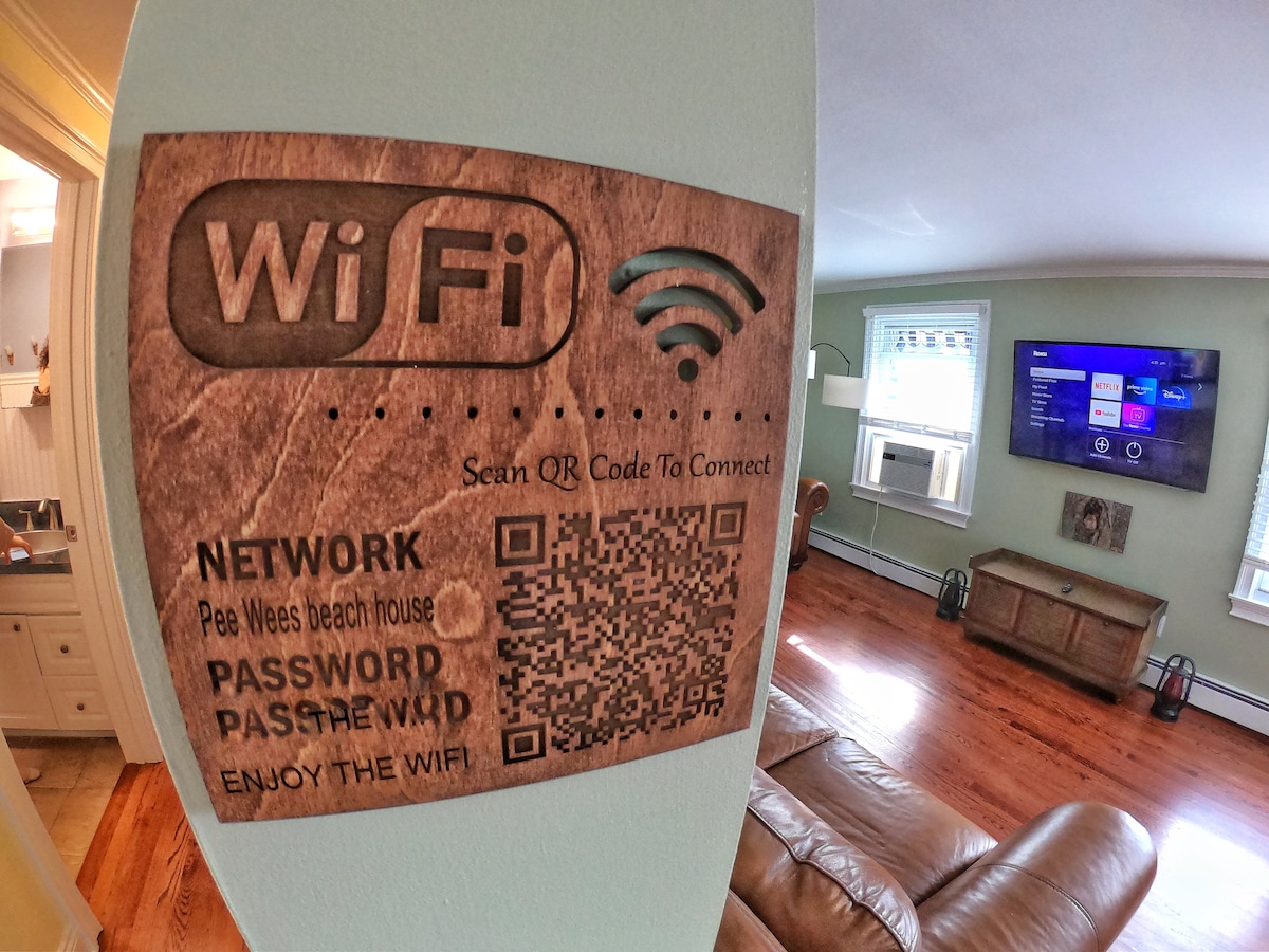A wall-mounted sign displays WiFi information, featuring a QR code for easy access. The surrounding light blue wall complements the spacious living area, which includes a wooden cabinet and a flat-screen television. Natural light enters through a nearby window, creating a welcoming environment.