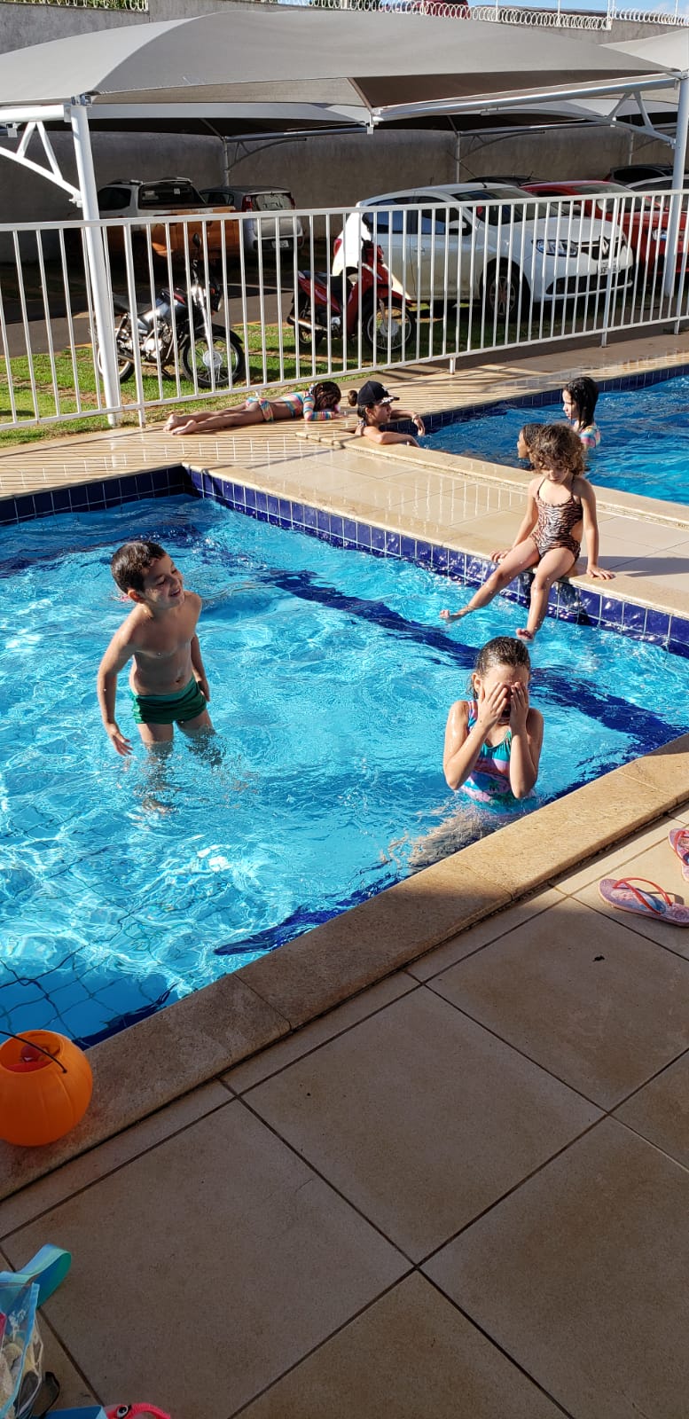 A clear swimming pool is visible, with children playing in the water and sitting along the edge. Sunlight reflects on the water's surface, while poolside tiles provide a spacious area for relaxation. A fence separates the pool from the surrounding area.
