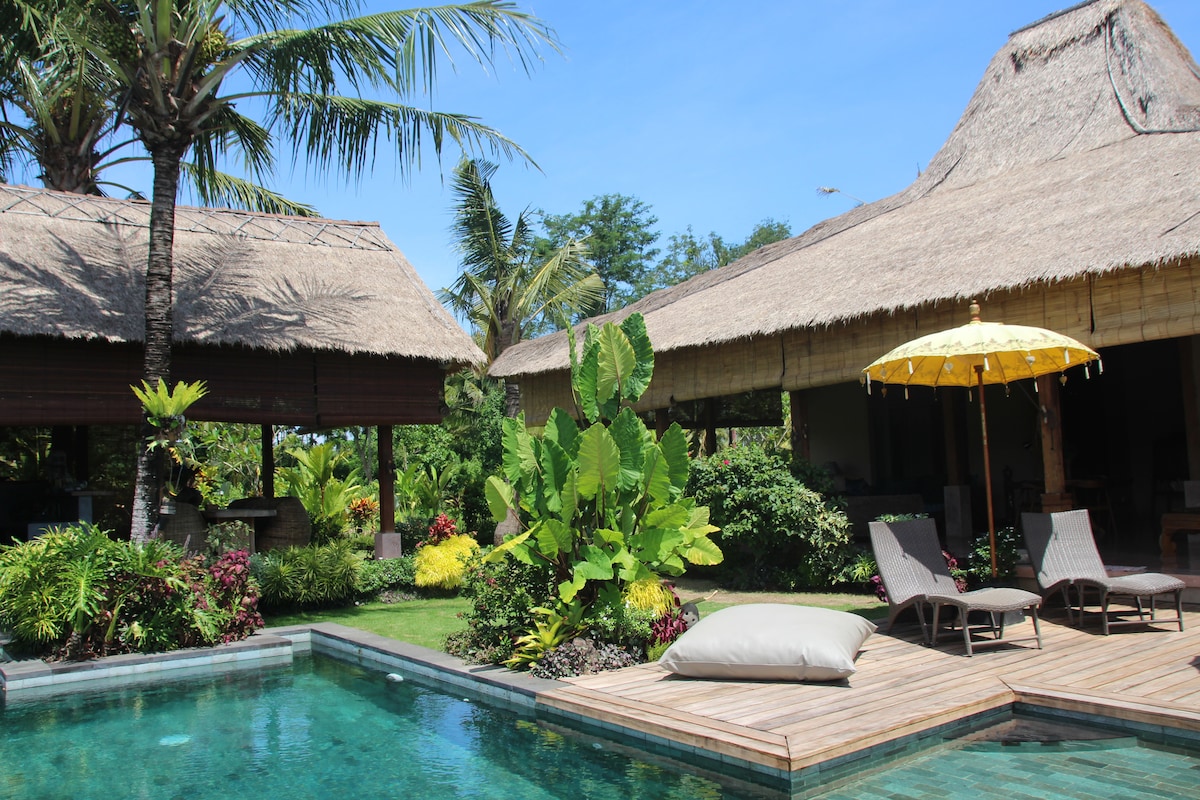 A private villa features a tranquil infinity pool surrounded by lush greenery, with lounge chairs positioned on a wooden deck. Thatched roofs adorn the nearby buildings, complemented by a yellow umbrella providing shade. Tropical plants enhance the serene atmosphere.