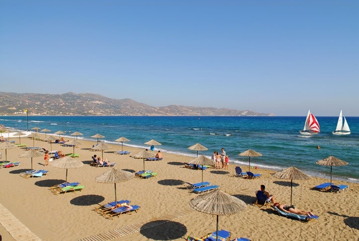 A sandy beach stretches along the coastline, lined with numerous umbrellas and lounge chairs for relaxation. The clear blue water gently laps at the shore, while sailboats glide across the horizon under a clear sky, showcasing a serene and inviting seaside atmosphere.