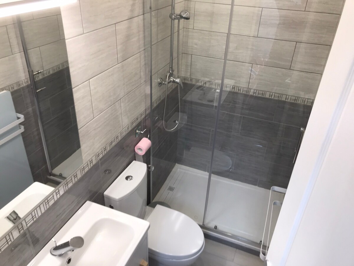 A modern bathroom featuring a walk-in shower enclosed by glass panels. The minimalist design includes a sleek sink and toilet, complemented by light grey tiles. A large mirror reflects the space, while a pink towel adds a touch of color to the neutral tones.
