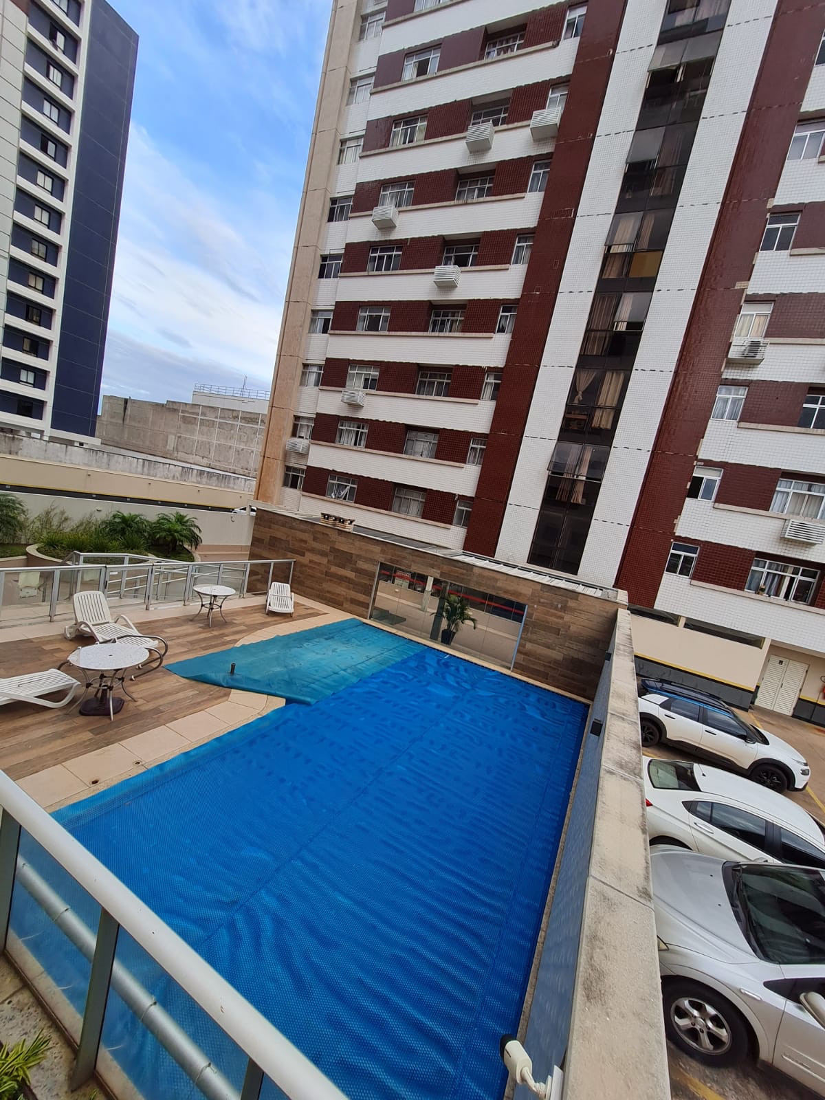 An outdoor pool area is visible with a blue cover. Surrounding the pool are several lounge chairs positioned on a wooden deck, offering a relaxing space. A multi-story building stands nearby, providing a backdrop of urban architecture.