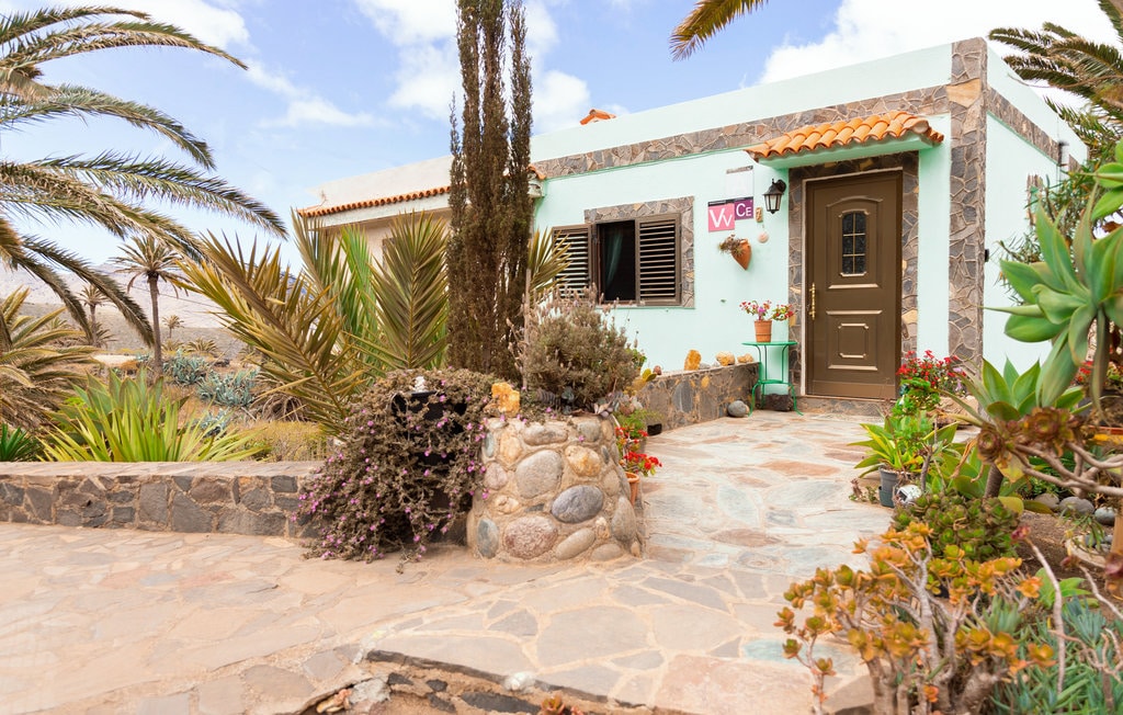 The entrance of a charming cottage is framed by vibrant greenery and desert plants. A stone pathway leads to a wooden door, complemented by window shutters. Colorful flowers and potted plants enhance the welcoming appearance of the exterior.