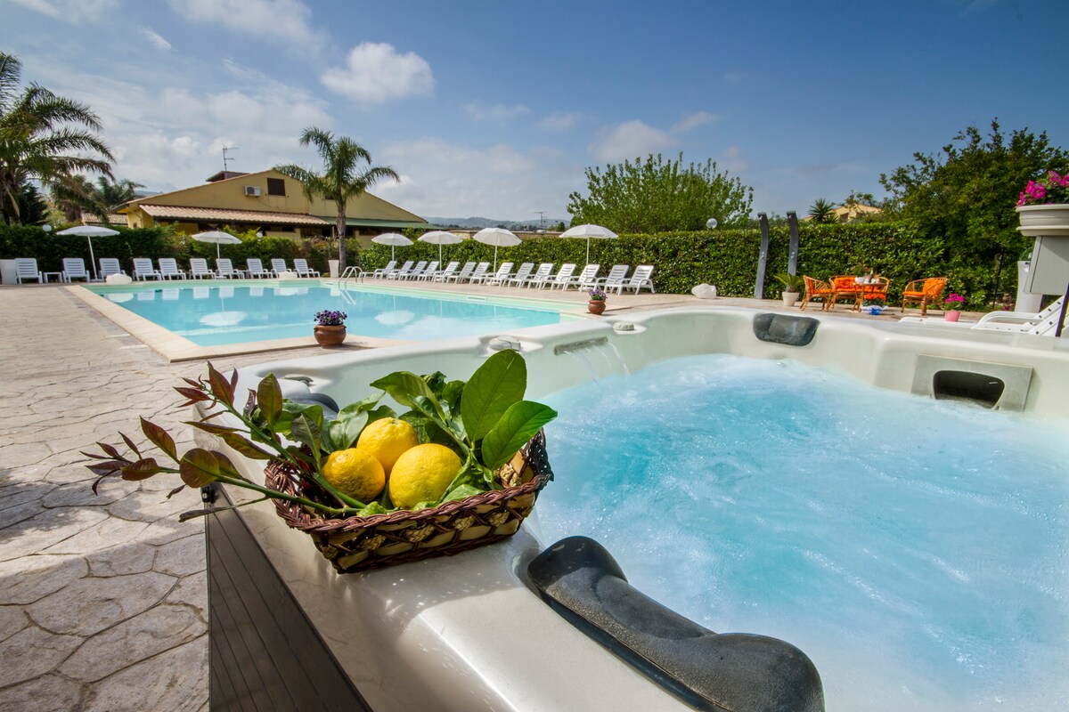 A spacious outdoor area is visible, featuring a large swimming pool and a jacuzzi tub. Surrounding the pool, sun loungers are arranged for relaxation. A decorative basket filled with lemons and greenery is placed in the foreground, enhancing the inviting atmosphere of the setting.