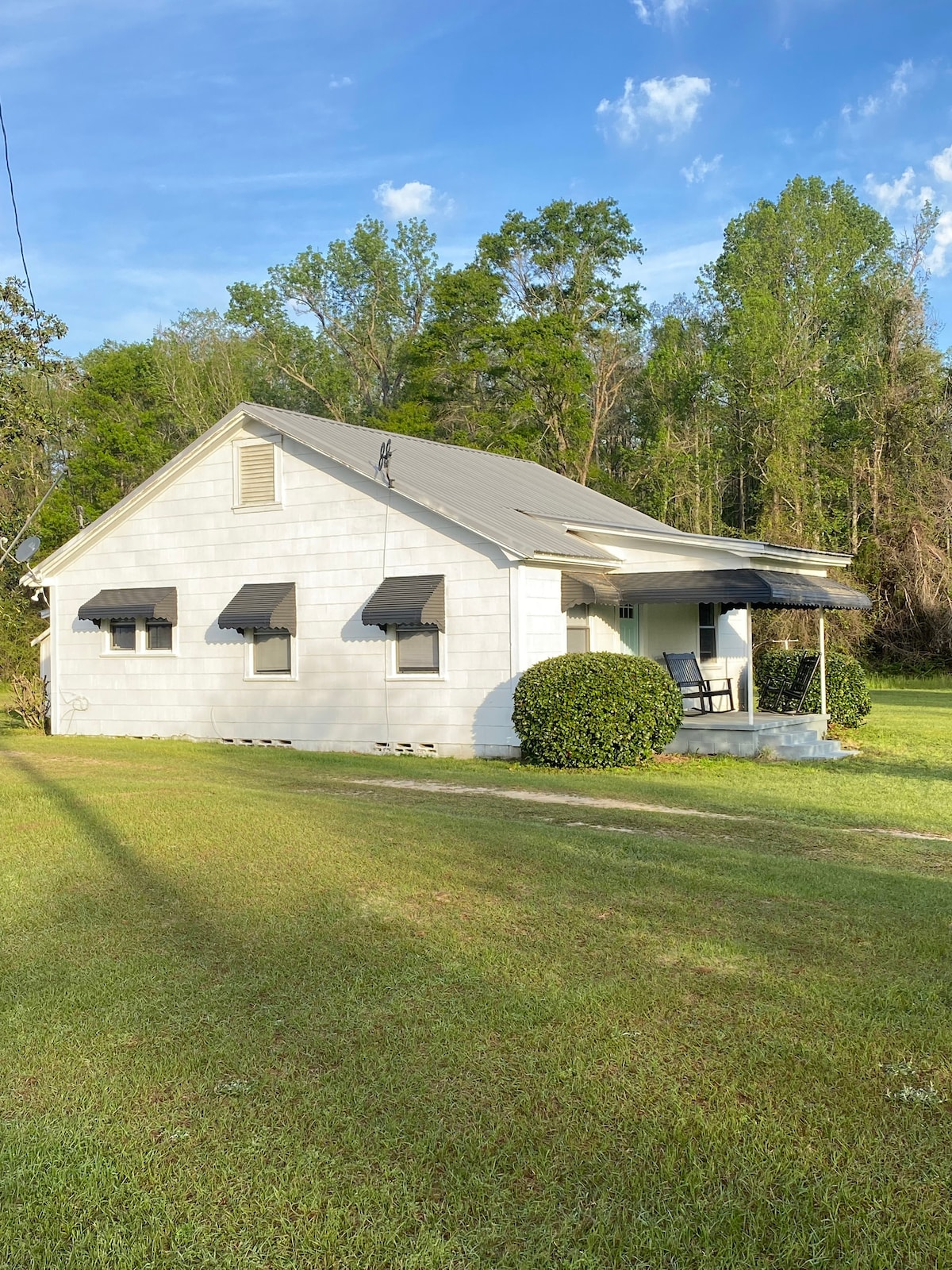 A charming bungalow is surrounded by lush greenery, featuring a sloped roof and black awnings over the windows. A covered porch with a rocking chair invites relaxation. The well-maintained lawn extends in front, offering ample space for outdoor enjoyment.
