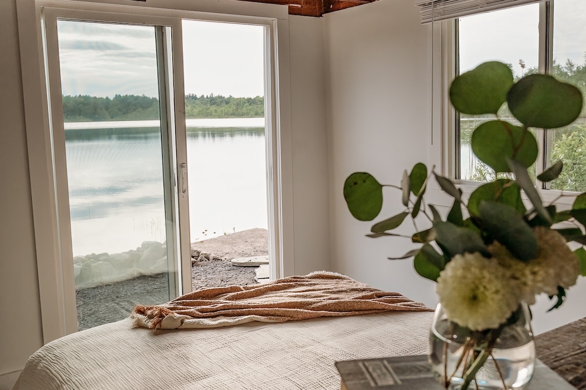 A cozy bedroom features a spacious bed adorned with a soft blanket. Large sliding doors reveal serene views of the lake, allowing natural light to fill the space. A vase with fresh greenery and flowers adds a touch of nature to the tranquil setting.