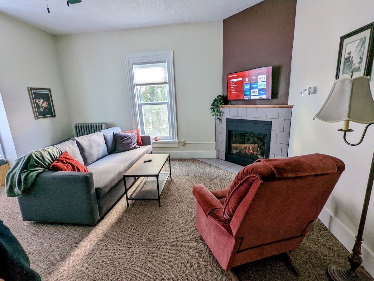 A comfortable living area is presented with a gray sofa and a burgundy armchair, arranged around a simple coffee table. A flat-screen television is mounted on the wall beside a marble fireplace. Natural light filters through the window, illuminating the warm, inviting space.