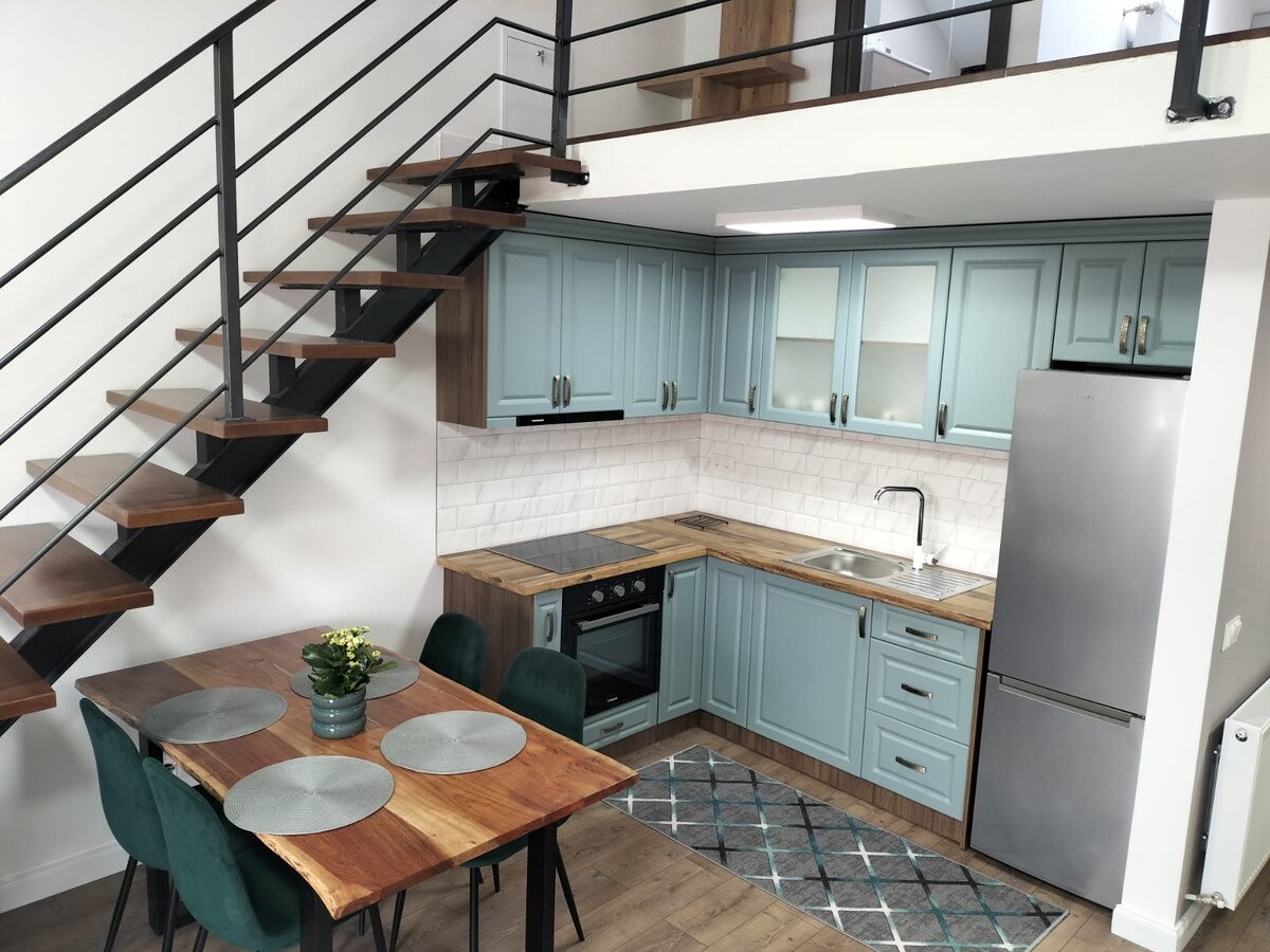 A modern kitchen area is featured, showcasing light blue cabinetry and a wooden countertop. Stainless steel appliances are present, including a refrigerator and oven. A dining table with green upholstered chairs is positioned nearby. A staircase leads to the upper level, enhancing the open layout.