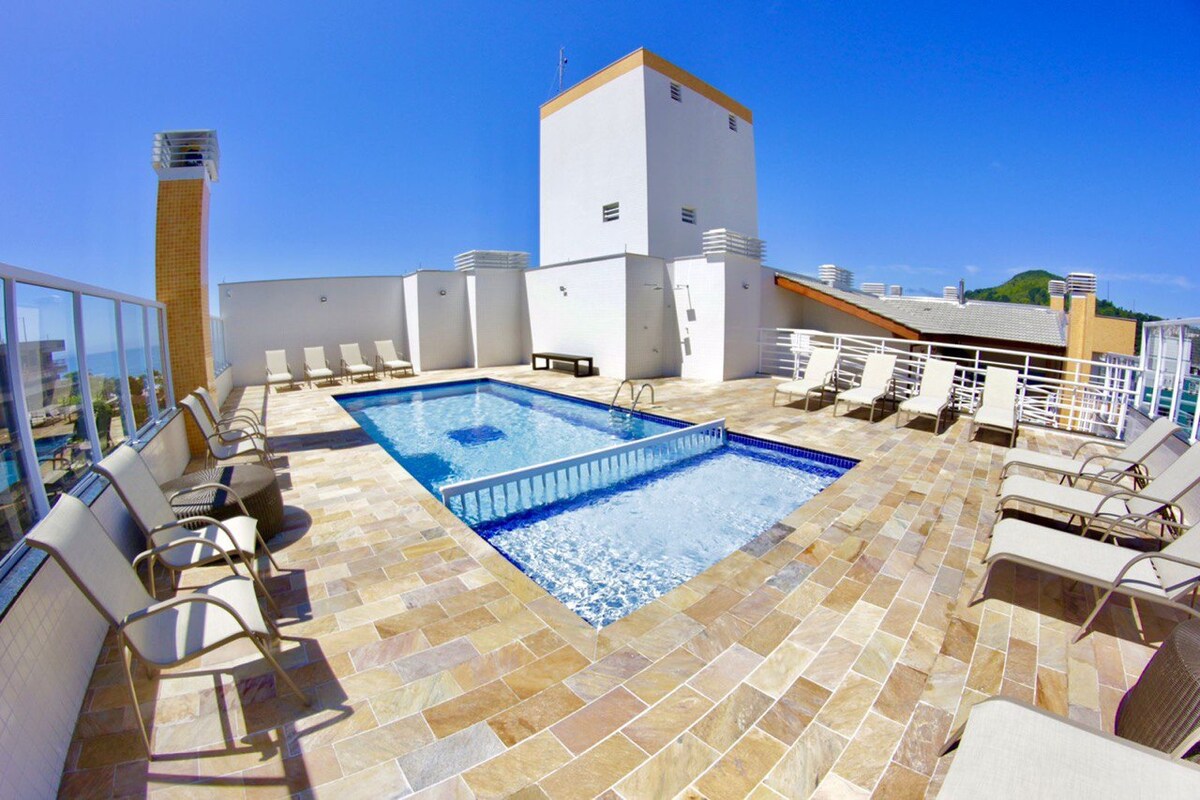 A rooftop pool area is featured, surrounded by lounge chairs and an expansive blue sky above. The pool is designed with a shallow section and tile detailing, offering a refreshing spot for relaxation. Neutral-colored stones cover the flooring, enhancing the outdoor atmosphere.
