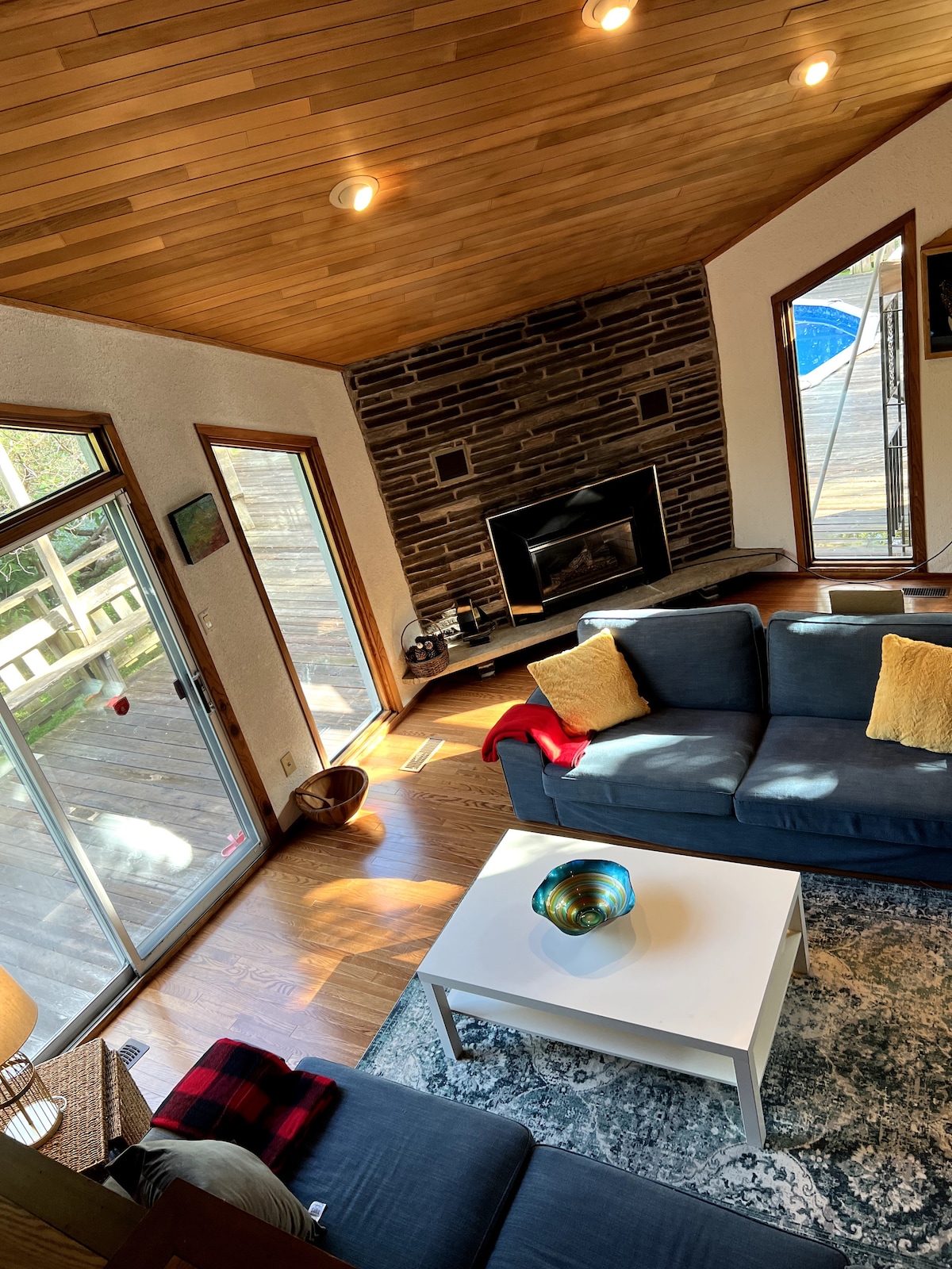 A spacious living area features large windows allowing natural light to illuminate the space. A stone fireplace anchors the room, while two sofas and a white coffee table create a comfortable seating arrangement. Decorative cushions add a touch of color to the seating.