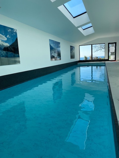 An indoor pool is seen, featuring clear turquoise water reflecting overhead skylights. Large windows on one side offer a view of the gardens and sea, while artwork adorns the walls, enhancing the serene atmosphere of the space.