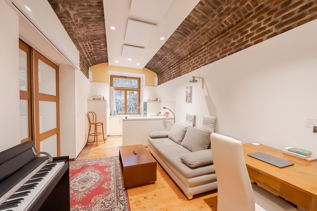 An inviting living space features a spacious gray sofa positioned in front of a sleek piano. Natural light filters through a large window overlooking greenery, while a rustic ceiling with exposed brick adds character. A work desk and modern kitchen area are also visible in the layout.