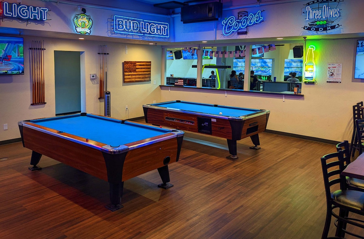 A recreational space features two pool tables with bright blue felt, flanked by colorful illuminated signs on the walls. A large mirror reflects the area, and wooden chairs are positioned around a low table, adding to the social atmosphere.