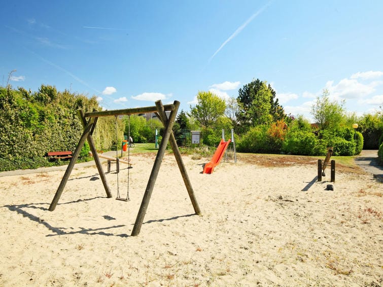 A sunny playground area features a wooden swing set and a slide, both situated on sand. A few benches and greenery line the perimeter, providing a natural setting. The clear blue sky enhances the bright atmosphere, inviting families to enjoy outdoor activities.