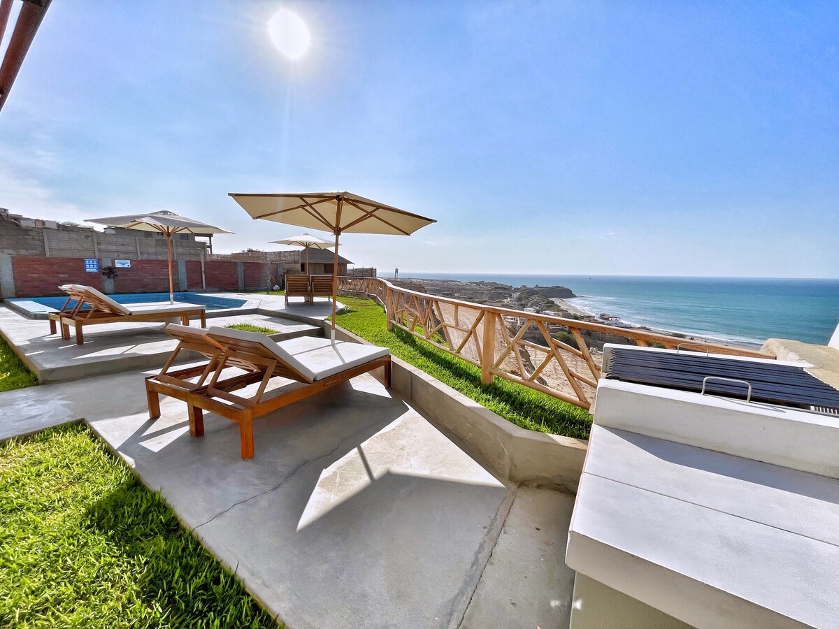 A sunlit area features loungers placed on grass, accompanied by umbrellas for shade. The pool is visible alongside a breathtaking view of the blue ocean and coastline, with the sun shining brightly overhead.