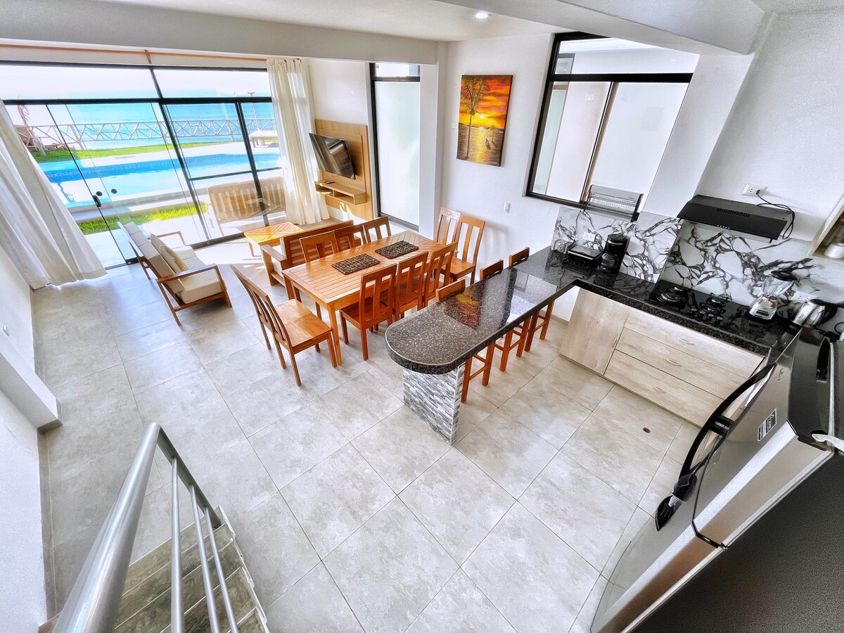 A bright open space is observed, featuring a dining area with a large table and wooden chairs. A modern kitchen with sleek countertops is visible, complemented by expansive glass doors that provide a view of the pool and ocean beyond.