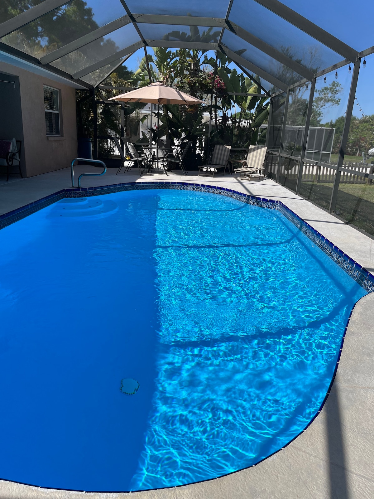 A private pool area is enclosed by a screened-in lanai, featuring clear blue water reflecting sunlight. A patio umbrella and several chairs are positioned nearby, surrounded by lush greenery, creating a relaxing outdoor space.