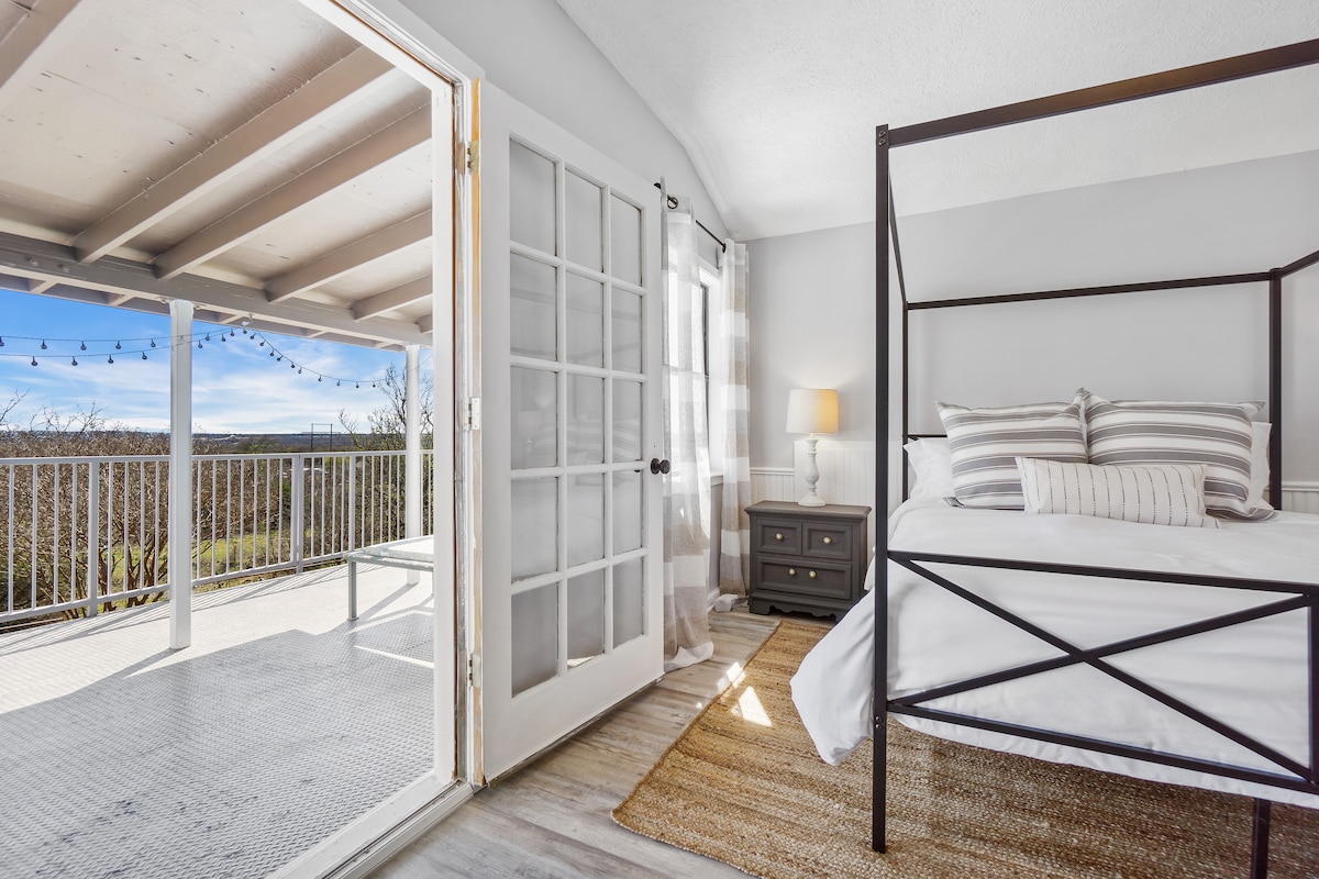A bright sleeping area features a canopy bed with soft gray and white bedding. Light pours in through French doors that lead to a spacious balcony, showcasing a scenic view across the landscape. A nightstand stands beside the bed, complementing the neutral decor.