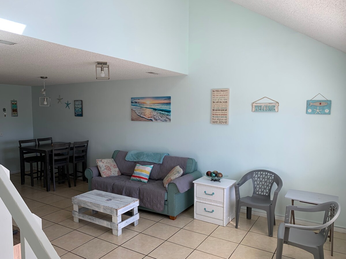 The living area features a light blue wall that enhances the spacious feel. A comfortable sofa is positioned alongside a white coffee table, while a dining table with chairs is located in the background. Various decorative elements, including wall art, accent the space.