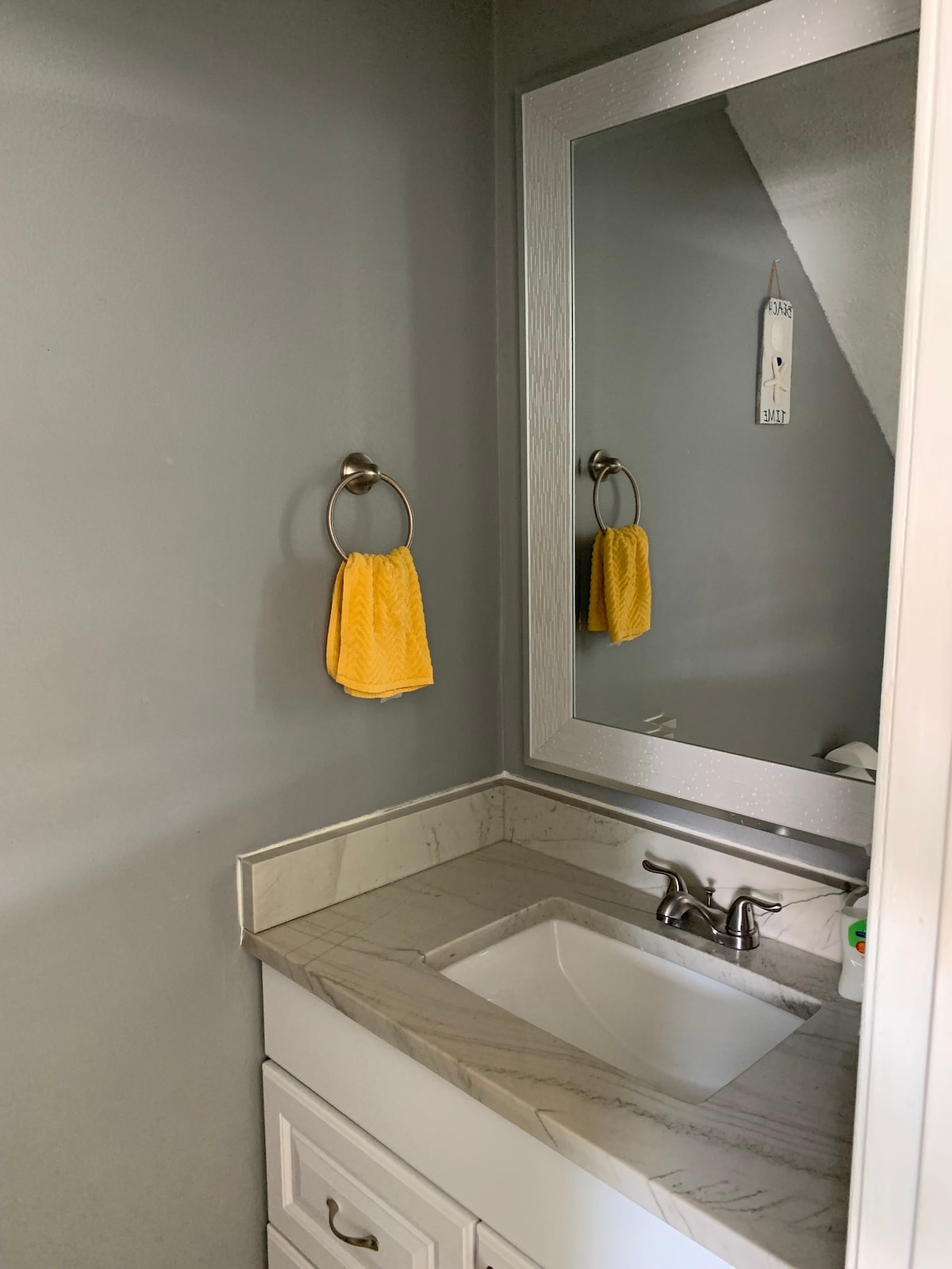 A bathroom features gray walls and a simple design, highlighting a light-colored countertop with a sink. A large mirror reflects the space, and a yellow towel hangs from a metal ring. The layout is functional and tidy, providing essential amenities.