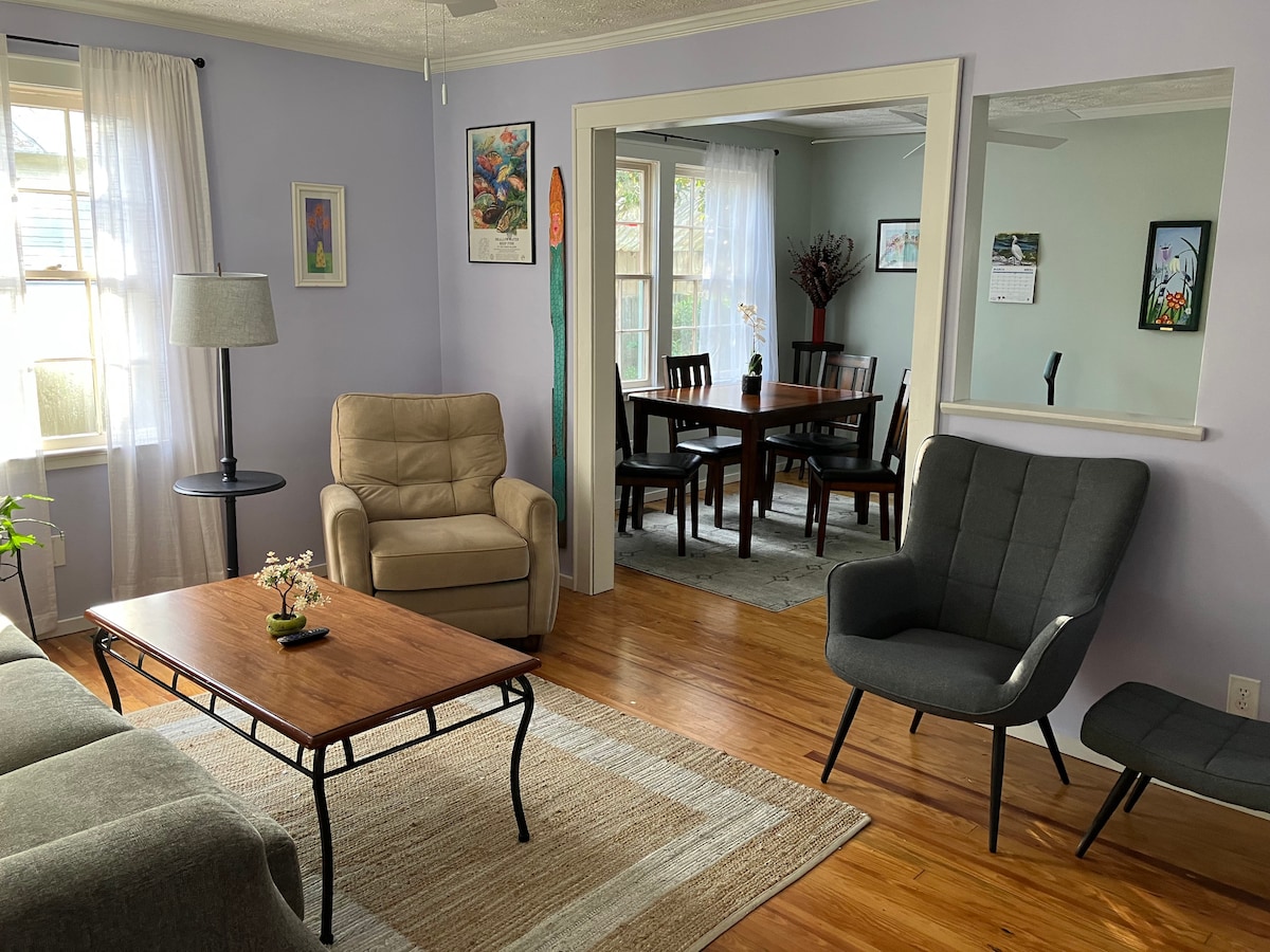 A cozy living room is visible, featuring a light brown sofa, a matching chair, and a dark gray chair. A wooden coffee table sits in the center on a light area rug. Natural light enters through large windows, creating a bright and inviting space.