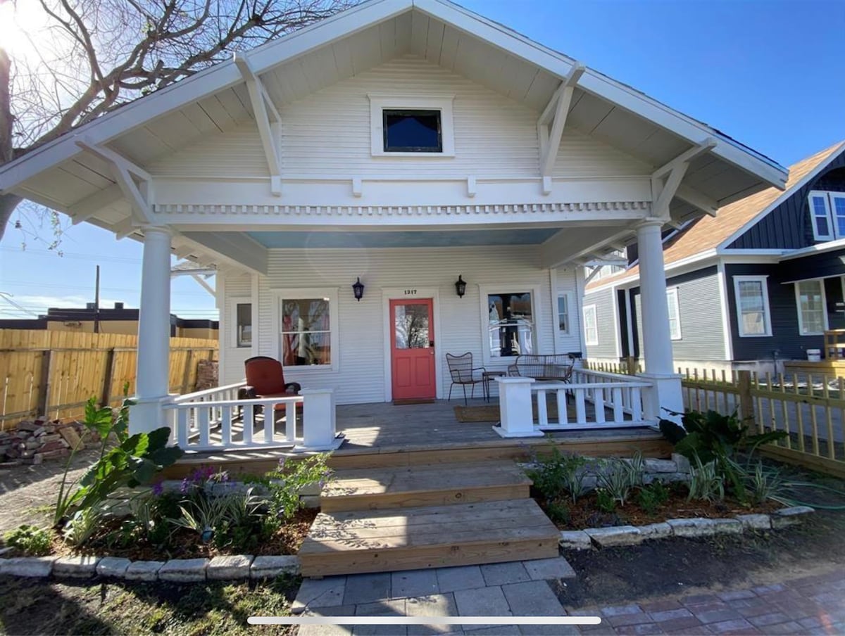 A charming front porch features a wooden deck with seating arrangements, including two chairs and a small table. The house's white facade is complemented by a bright red door. Lush plants border the porch, adding a touch of greenery and inviting ambiance.