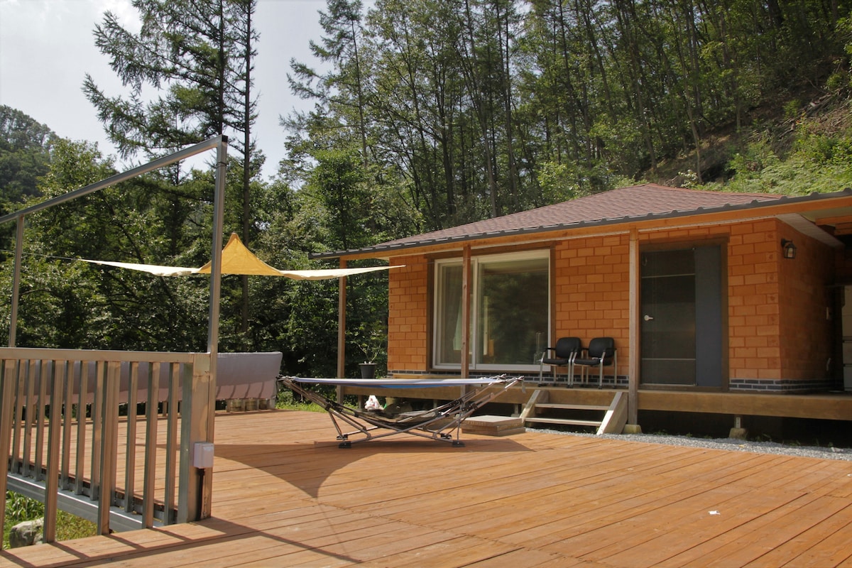 A cabin constructed with natural materials is set amidst a green forest backdrop. A spacious wooden deck features a lounge area with seating, and a sunshade is positioned overhead. Large glass doors provide access to the interior and allow natural light to enter.