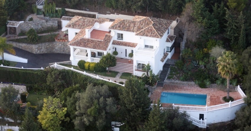 An elegant villa is surrounded by greenery, showcasing a well-manicured landscape. The property features a private pool adjacent to the outdoor terrace, with a distinctive tiled roof and multiple levels that enhance its architectural appeal.