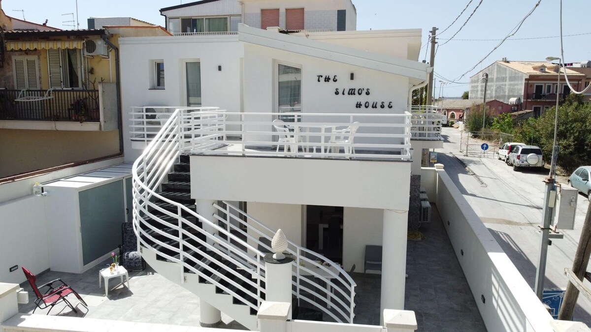 A modern building with a unique architectural design is displayed. A winding white staircase leads to an upper terrace, which is furnished with tables and chairs. The surrounding area features a quiet street and greenery in the background.