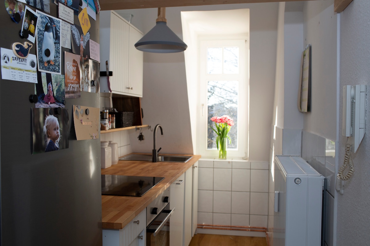 A compact kitchen area features wooden countertops and white cabinetry, creating a functional cooking space. A stainless steel fridge and a modern sink are visible. Sunlight streams through a window, highlighting a vase of flowers on the countertop, adding a touch of color to the room.