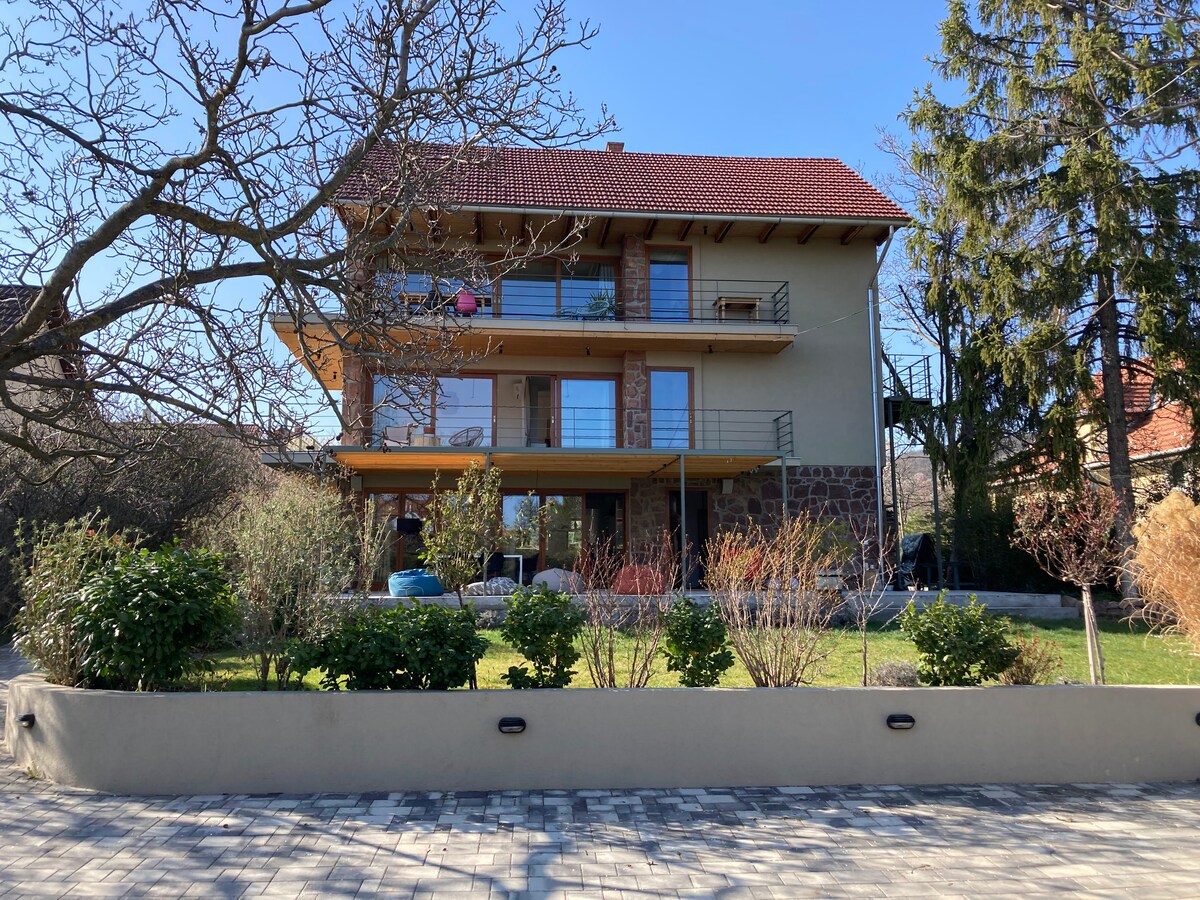 A three-story building with a wooden balcony and large glass doors is surrounded by well-maintained greenery. The front yard features a variety of plants and decorative elements, providing a natural setting. A clear blue sky forms the backdrop, enhancing the serene atmosphere.