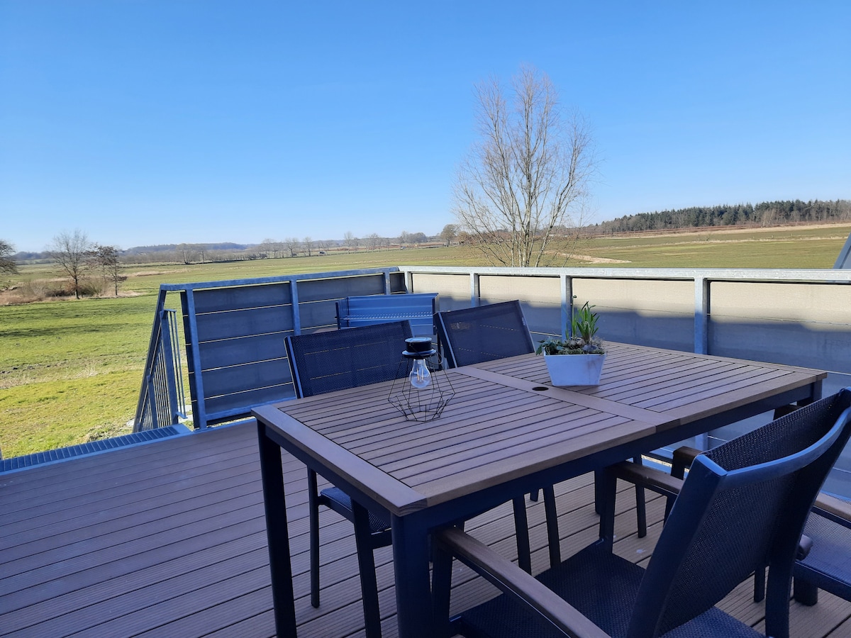 A spacious balcony features a wooden table surrounded by four dark chairs. A small potted plant rests on the table, accompanied by a decorative lantern. The view showcases expansive green fields under a clear blue sky.