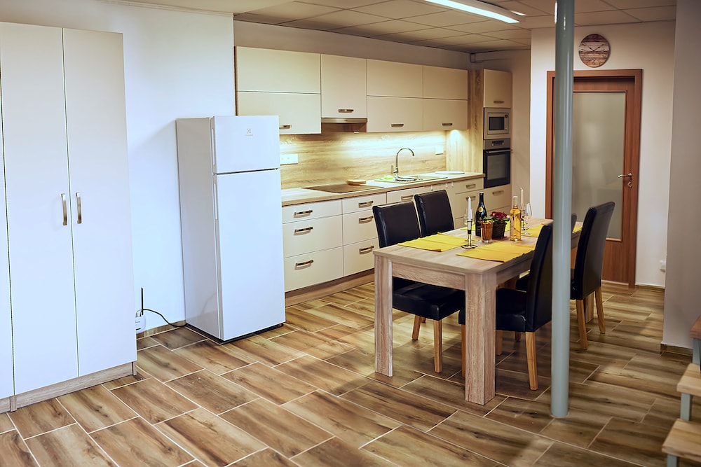 A modern kitchen and dining area are presented with light cabinetry and a central wooden dining table. Six chairs surround the table, while a white refrigerator and various appliances are visible, enhancing the fully equipped kitchen. Natural light fills the space through ample lighting.