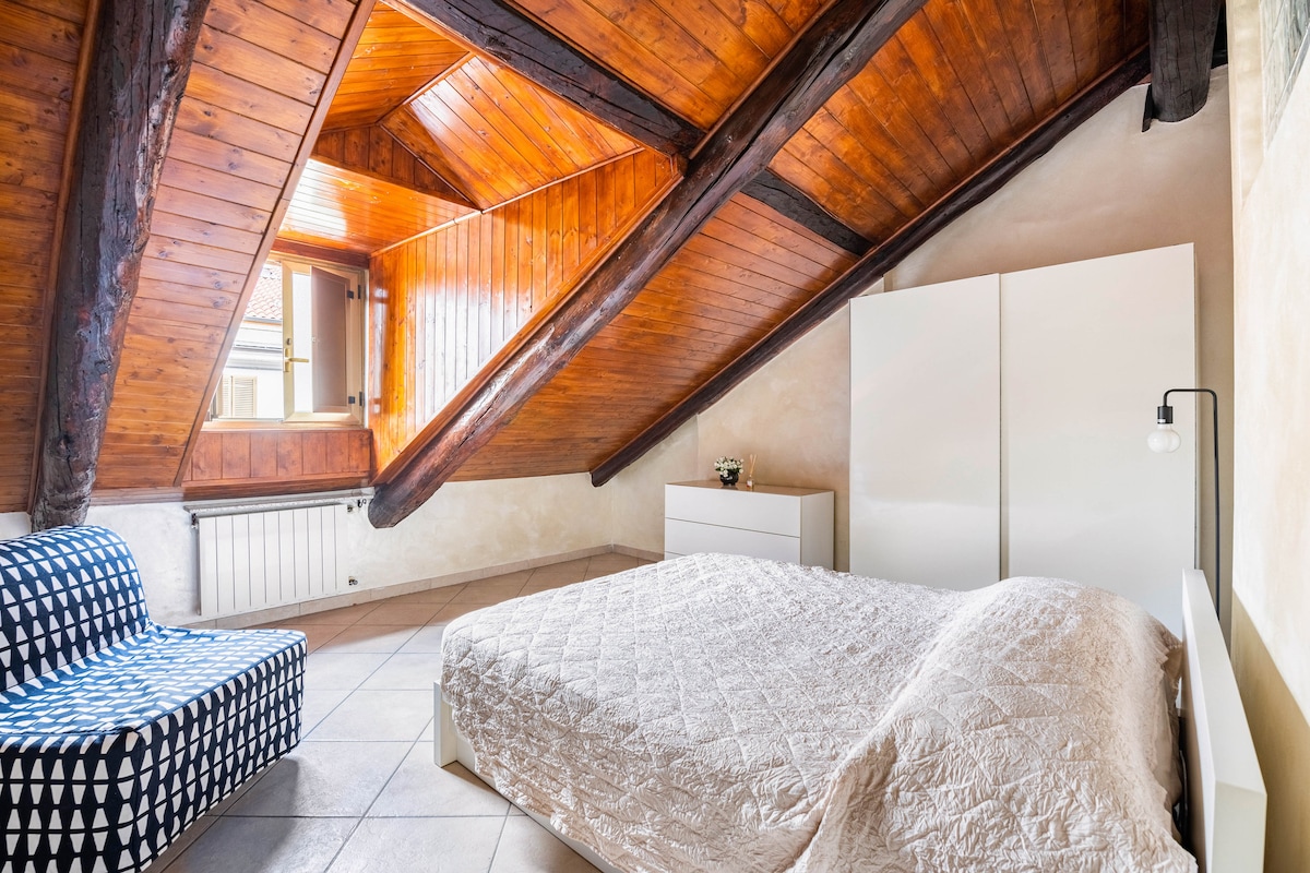 A cozy attic bedroom is showcased, featuring a spacious bed with a light quilt. A sleek white wardrobe stands against the wall, while a patterned chair adds a touch of contrast. Natural light filters through the window, illuminating the warm wooden beams.