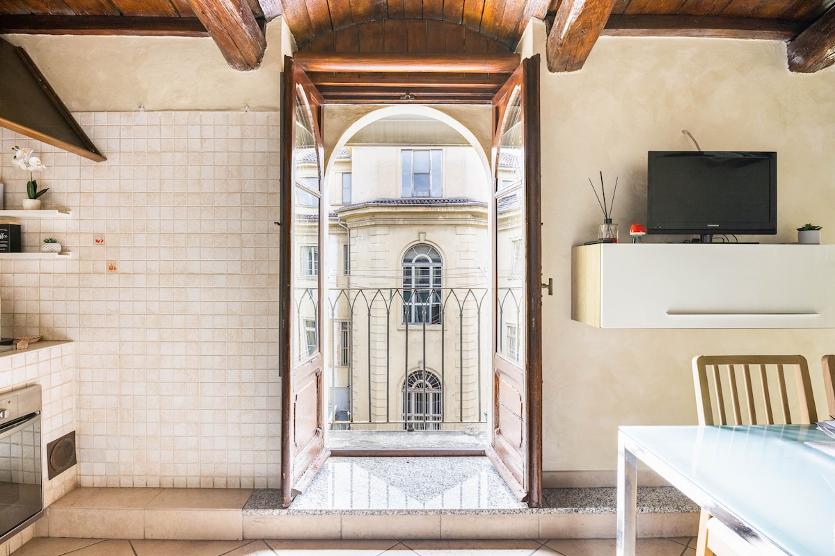 A spacious kitchen area is highlighted by arched wooden doors that open to a balcony, allowing natural light to flow in. The view includes a classic building facade, while a flat-screen TV is mounted on the wall. Wooden beams frame the room, adding character.