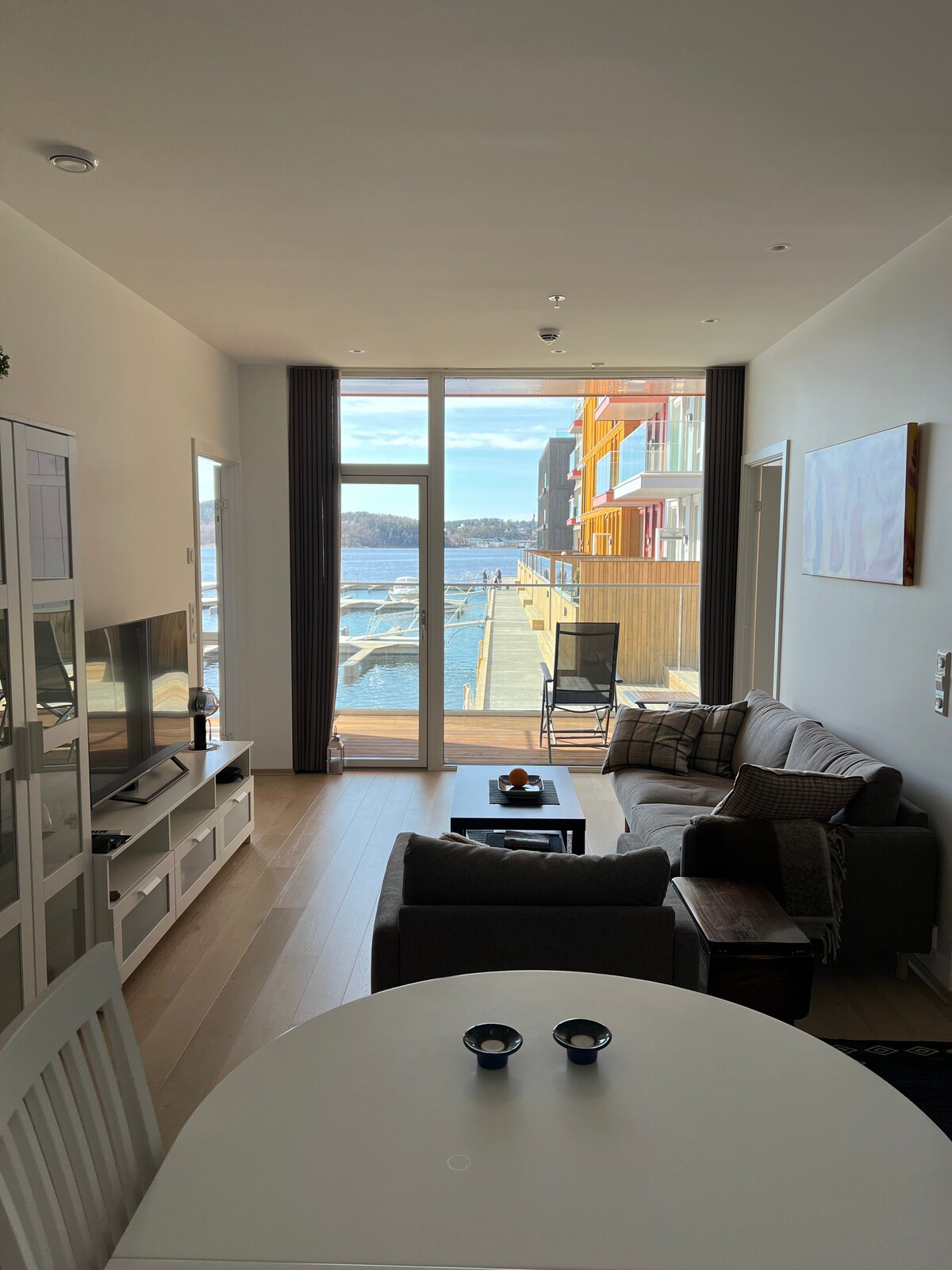 A combined living and dining area features large glass doors that open to a waterfront view. Natural light fills the space, highlighting a comfortable sofa and coffee table. A dining table is set for two, with additional seating visible in the cozy setting.
